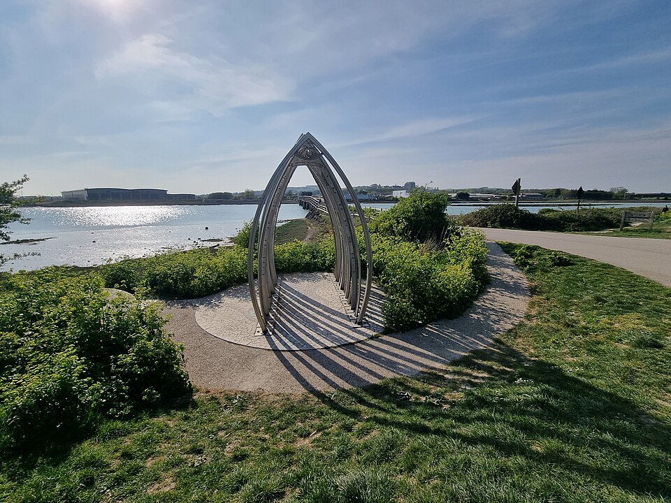 Image shows a silver metal memorial structure to victims of the 2015 Shoreham Airshow disaster by the River Adur during the daytime. The sun is casting a shadow behind the structure onto the grass