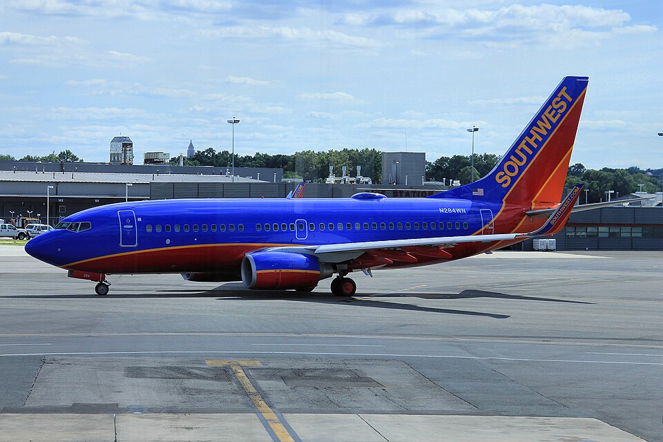 A Southwest Airlines Boeing 737-800 (registration N284WN) on the tarmac at an airport, featuring the airline's vibrant blue, red, and orange livery with "SOUTHWEST" prominently displayed on the tail, set against a background of airport terminal buildings, a control tower, and a distant city skyline under a partly cloudy blue sky.