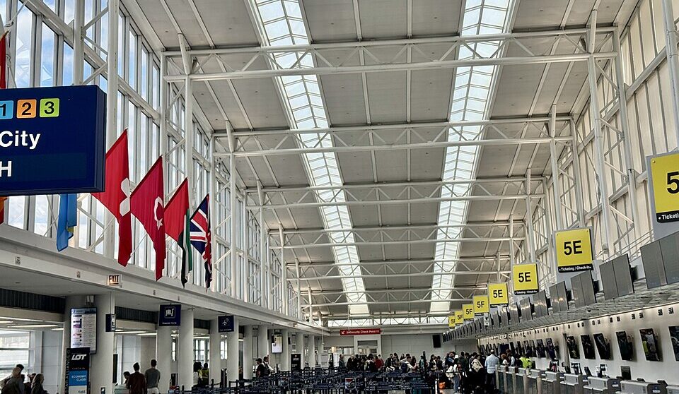 The interior of a large, modern airport terminal check-in hall with a high, white steel truss ceiling and skylights. Rows of check-in counters with yellow zone signs (5E, 5F) line the right side, where travellers are queued. On the left, large windows illuminate international flags, including Swiss and Turkish flags, hanging from an upper level.