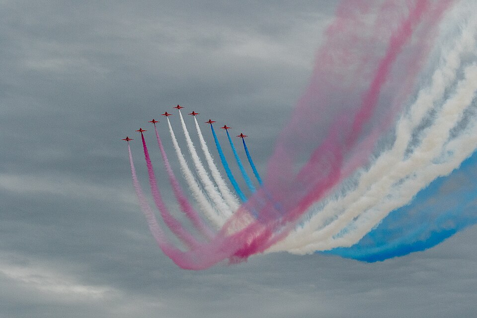 Image shows 9 aircraft from the Red Arrow display team with red, white and blue plumes trailing behind, on a cloudy day in 2015 at Clacton Air Show