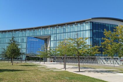 A modern, multi-story office building with a sleek glass and metal facade bearing the "AIRBUS" logo above the main entrance, surrounded by green lawns, young trees, and a concrete walkway under a clear blue sky.