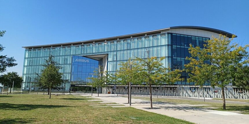 A modern, multi-story office building with a sleek glass and metal facade bearing the "AIRBUS" logo above the main entrance, surrounded by green lawns, young trees, and a concrete walkway under a clear blue sky.