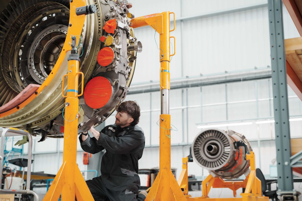  AerFin engine with bright orange stabilisers hoisting it up in the air. A maintenance technician is diligently working on the engine from below. Another engine can be seen in the background.