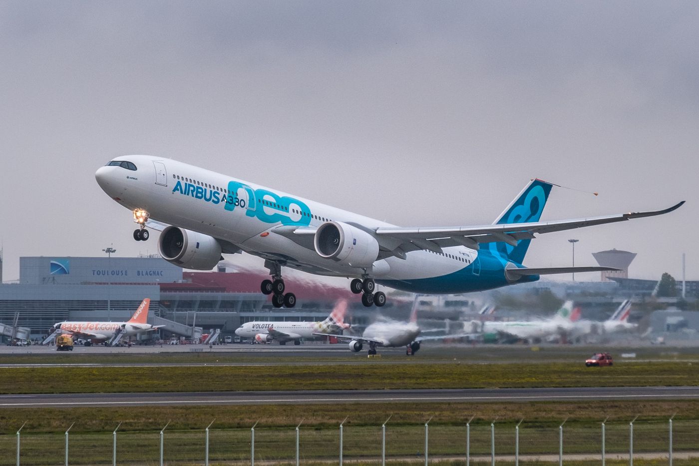 An Airbus A330-900neo aircraft in white and blue taking off with a cloudy sky backdrop and grass surrounding the runway