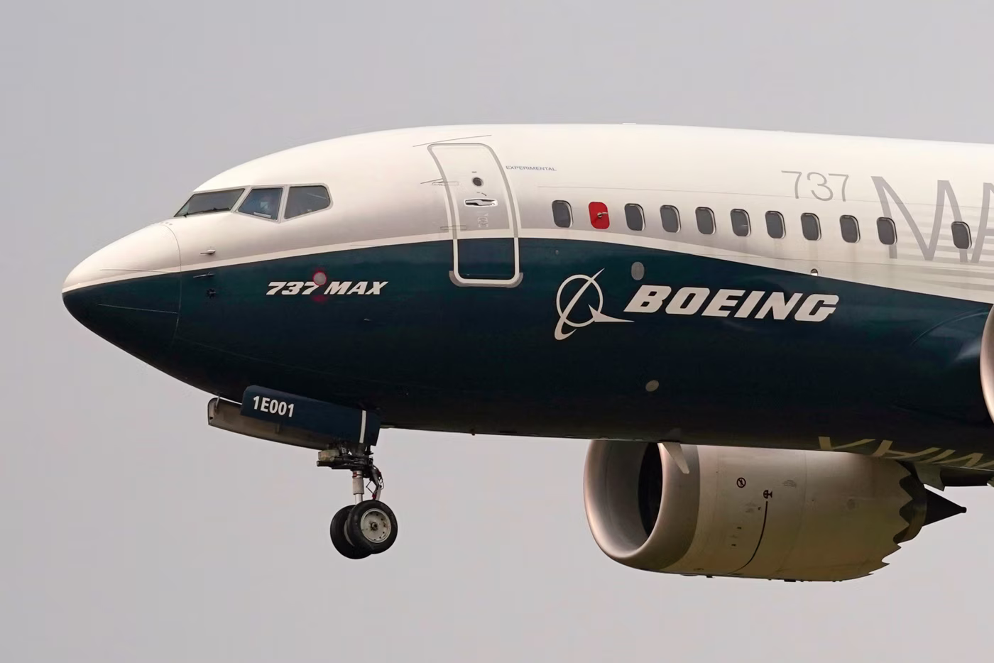 The nose gear and nose cone of a Boeing 737 MAX aircraft flying through a grey sky, with its landing gear out.