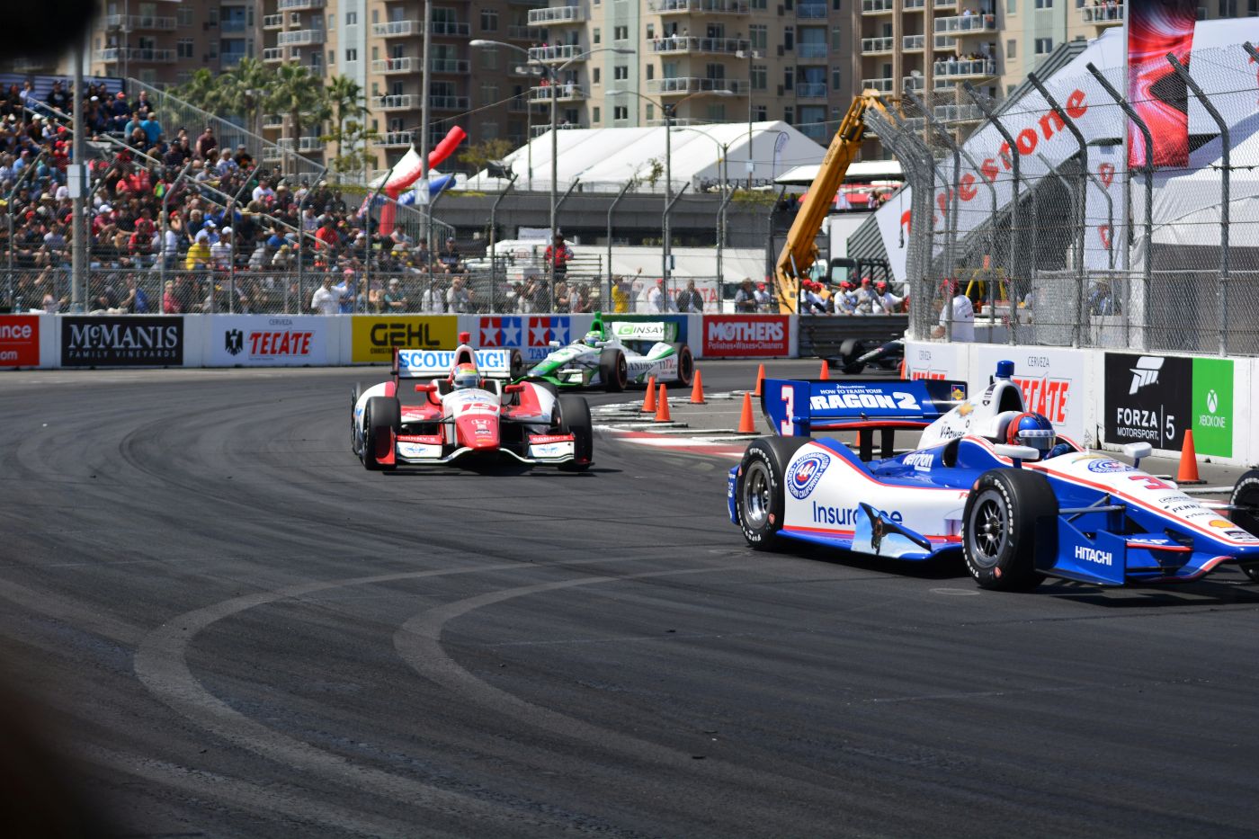 A picture of race cars swerving around a racecourse corner during a grand prix event with crowds cheering in the stands. The cars are blue and white, and red and white.