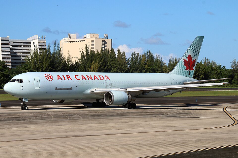 An Air Canada Cargo Boeing aircraft taxing on the runway. Buildings and blue sky in the background. White and red livery, with a maple leaf on the tail.