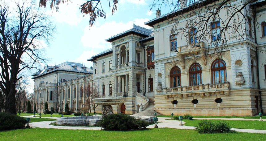 The exterior of the façade of Bucharest's Cotroceni Palace. Grass in the foreground, blue sky in the background. Fountain in front of the building.