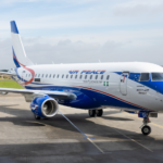 An Embraer E175 aircraft in blue and white sititng on tarmac with green grass and a cloudy sky in the background.