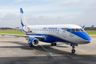 An Embraer E175 aircraft in blue and white sititng on tarmac with green grass and a cloudy sky in the background.