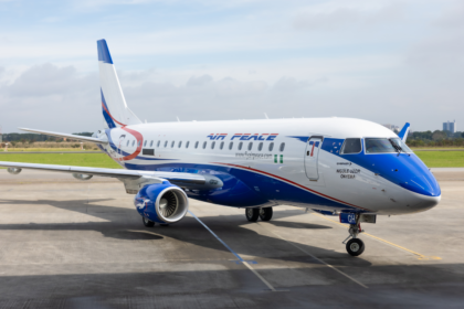 An Embraer E175 aircraft in blue and white sititng on tarmac with green grass and a cloudy sky in the background.