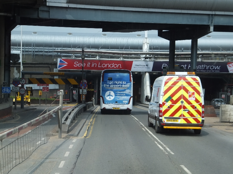 The entry of Terminal 2 car part at London Heathrow Airport. From the bottom to the middle of the image is tarmac road. In the middle of the image, one van and one coach can be seen driving into the car park, beneath a red sign that reads "See it in London." The top of the image shows what looks like the underside of a bridge or tunnel just before the grey roof of the car park's entrance. 