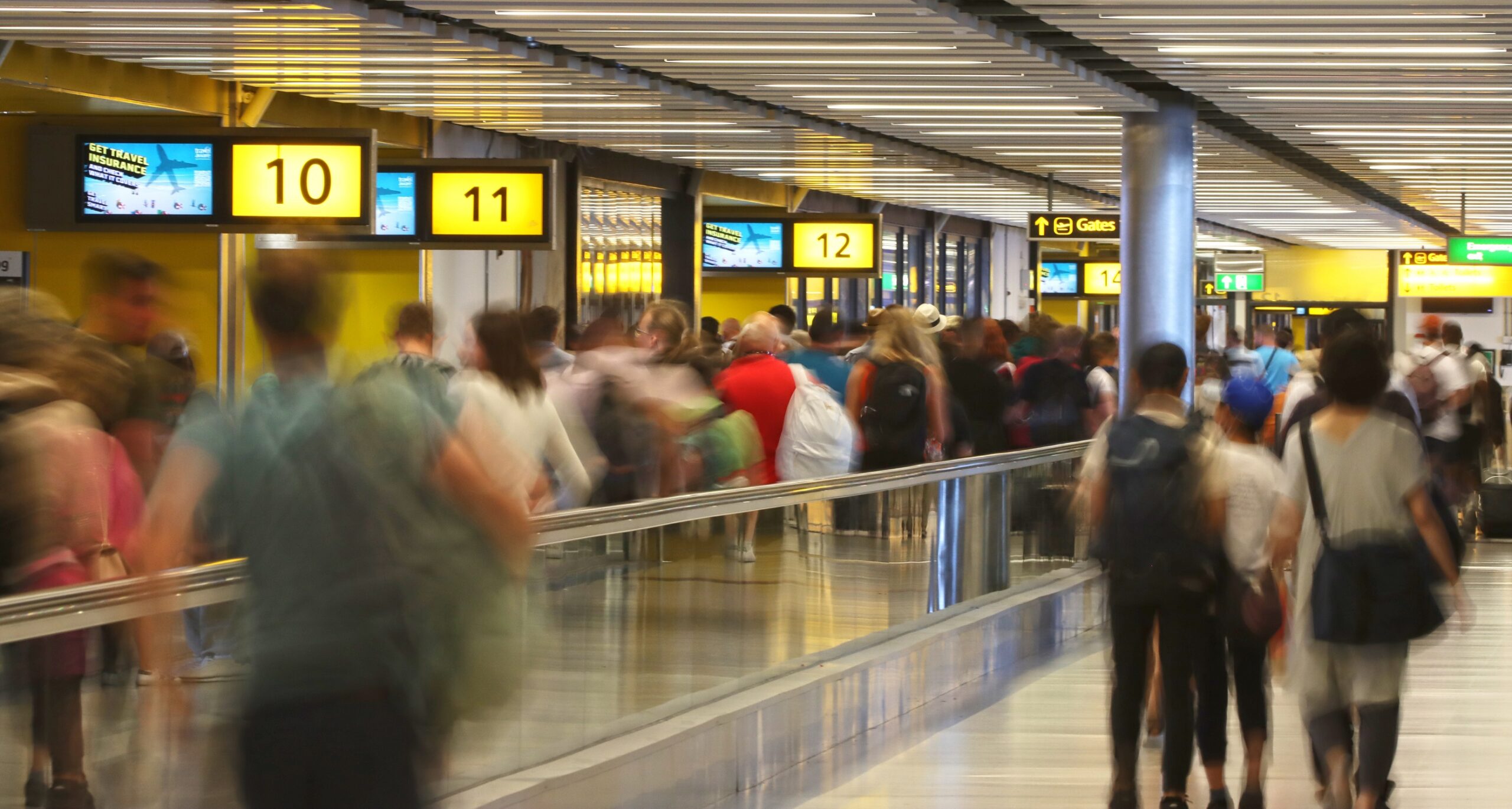 Passengers move through a busy airport departure area, passing gate signs and a moving walkway. Motion blur captures the flow of travellers making their way towards the gates