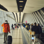 Travellers make their way through a bright airport walkway, some pulling suitcases, with a moving walkway on one side and large windows lining the terminal. The modern pier features curved ceiling panels and steel beams stretching into the distance