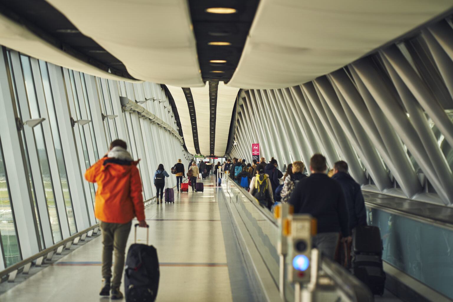 Travellers make their way through a bright airport walkway, some pulling suitcases, with a moving walkway on one side and large windows lining the terminal. The modern pier features curved ceiling panels and steel beams stretching into the distance