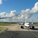 Planes queuing at a Heathrow Airport terminal, with one jet facing the camera. There is Grass on the sides and a blue sky at the backdrop
