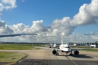 Planes queuing at a Heathrow Airport terminal, with one jet facing the camera. There is Grass on the sides and a blue sky at the backdrop