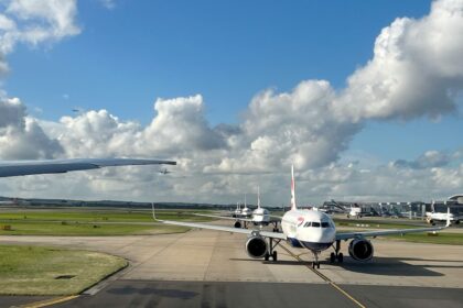 Planes queuing at a Heathrow Airport terminal, with one jet facing the camera. There is Grass on the sides and a blue sky at the backdrop