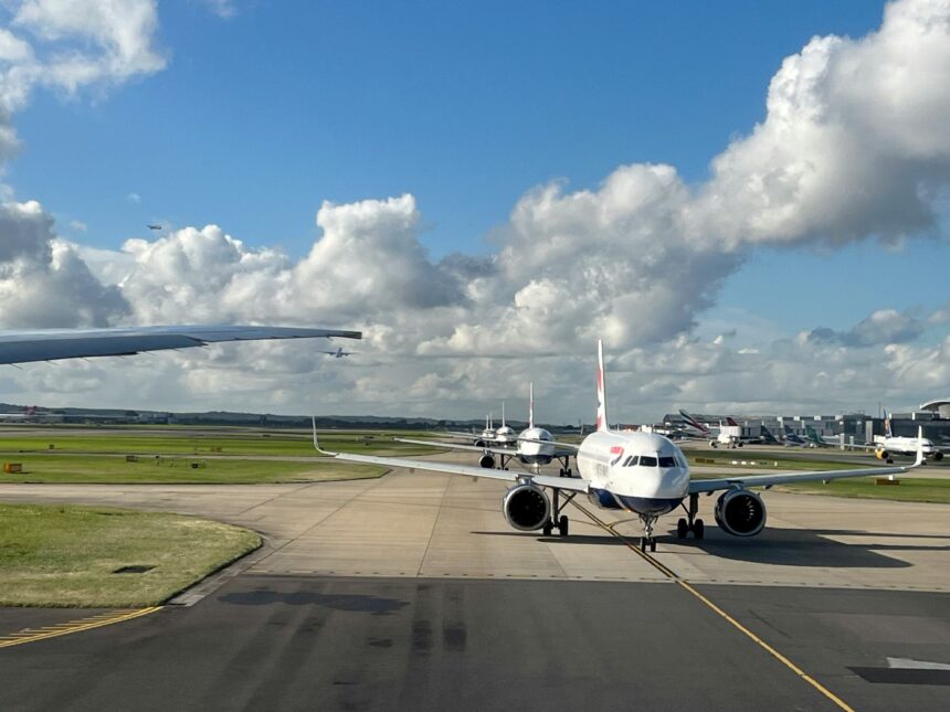 Planes queuing at a Heathrow Airport terminal, with one jet facing the camera. There is Grass on the sides and a blue sky at the backdrop