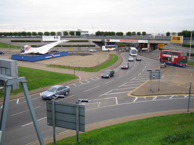 An image taken from above of a road leading to Heathrow Airport. The road emerges from the upper centre right and occupies most of the image, leading into the bottom left. In the bottom right corner a grass verge can be seen, opposite another grass verge on the central left. 5 cars and one coach drive towards the camera, and one car, one coach and one bus drive away. A bridge can be seen in the middle distance, just behind a statue of the Concorde on the central left, with large trees right in the background along the full length of the image. The top of the image shows a cloudy grey sky. 