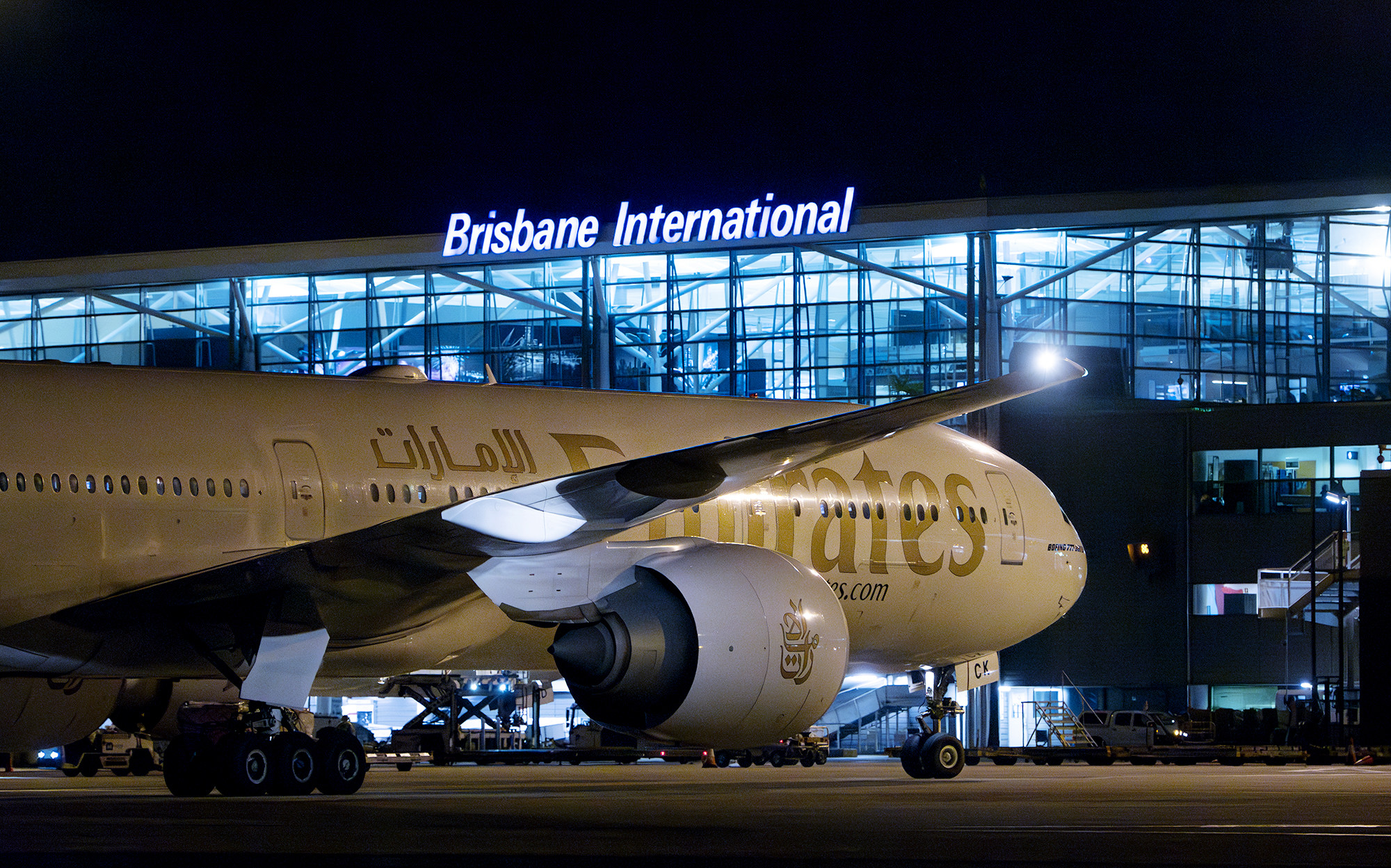 A white and gold Emirates jet parked at night in front of an airport terminal at Brisbane Airport (BNE).