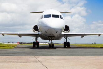 A head-on shot of an Alliance Airlines Embraer E190 sat on a runway in front of a grassy verge and a cloudy sky.