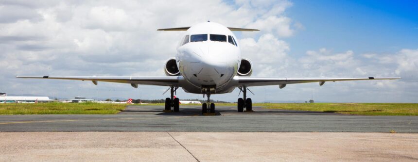A head-on shot of an Alliance Airlines Embraer E190 sat on a runway in front of a grassy verge and a cloudy sky.