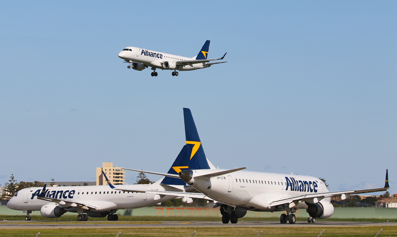 Two blue, white, and yellow Alliance Airlines jets sat on a runway. A third Alliance Airlines jet flies above them in front of a clear sky.