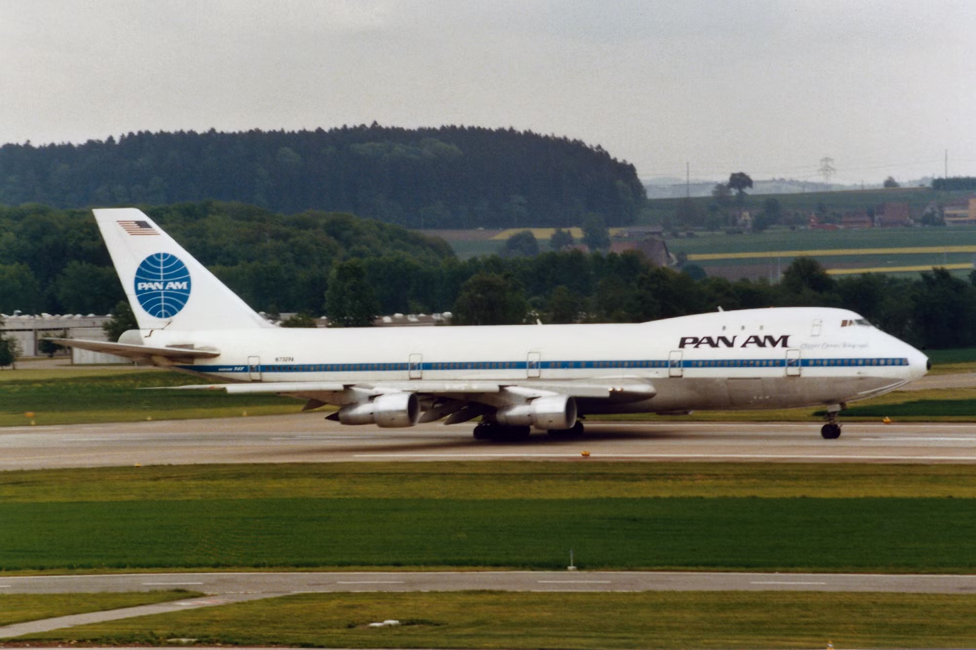 A blue and white Pan American World Airways jet sat on a runway behind a strip of grass, in front of a cloudy sky.
