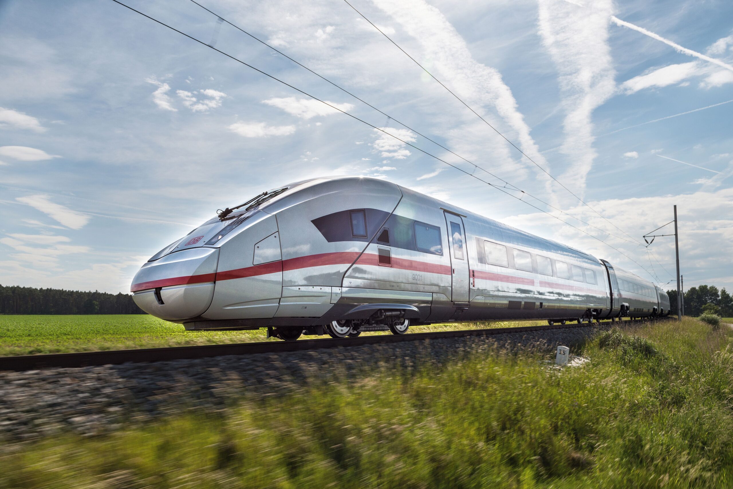 A red and silver Deutsche Bahn train travelling through a light green field on a sunny day.