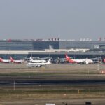Red and white planes sat on a runway in front of Shanghai Hongqiao International Airport (SHA). There is a grey sky behind them.