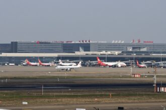 Red and white planes sat on a runway in front of Shanghai Hongqiao International Airport (SHA). There is a grey sky behind them.
