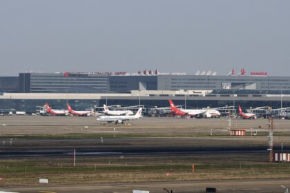 Red and white planes sat on a runway in front of Shanghai Hongqiao International Airport (SHA). There is a grey sky behind them.