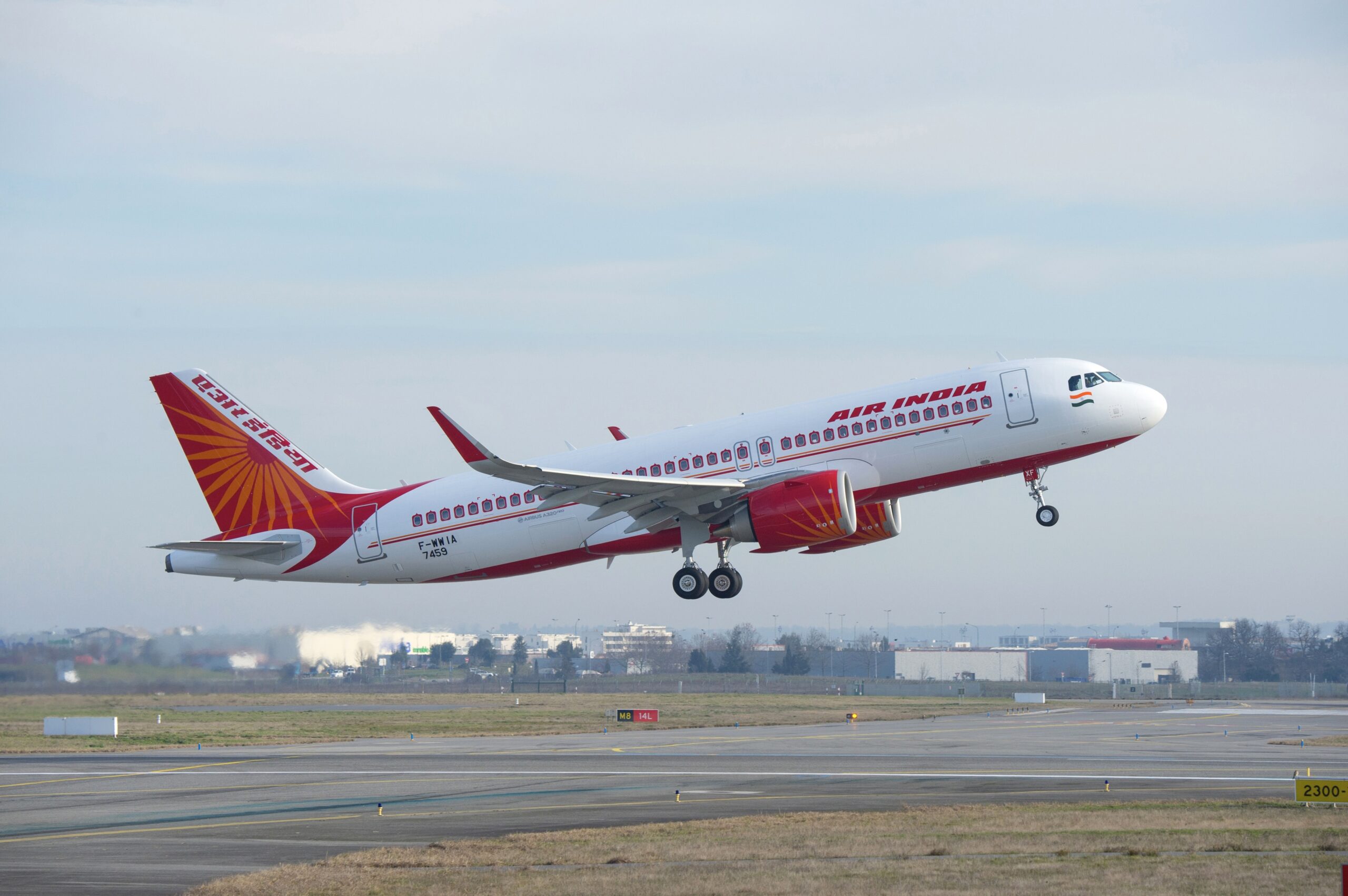 A red and white Airbus jet with an Air India livery flying upwards from an airport runway with buildings in the background.