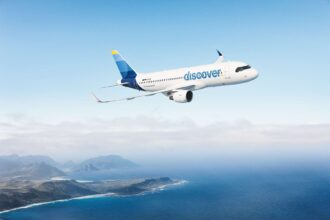 A blue and white Discover Airlines jet flying across an open sea with a green coastline to the bottom left of the image. The sky is blue but cloudy.