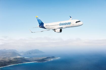 A blue and white Discover Airlines jet flying across an open sea with a green coastline to the bottom left of the image. The sky is blue but cloudy.
