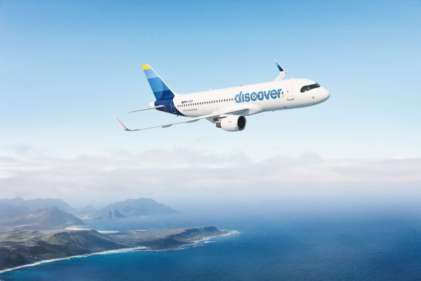 A blue and white Discover Airlines jet flying across an open sea with a green coastline to the bottom left of the image. The sky is blue but cloudy.