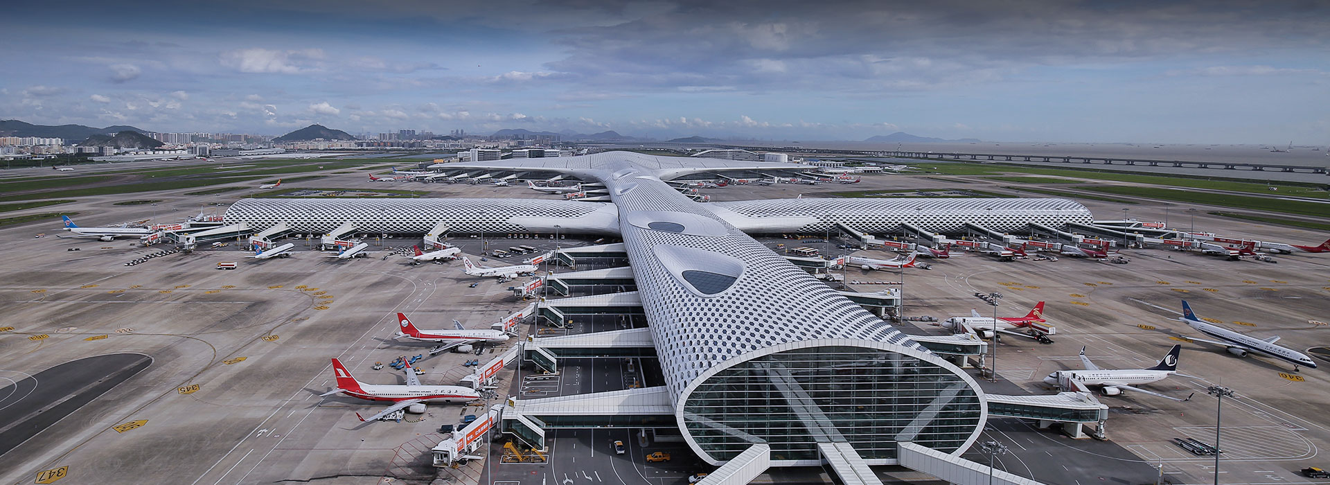 Shenzhen Bao’an International Airport (SCZ). A metal and glass airport terminal is flanked by white planes parked on a grey runway. There is a cloudy sky behind.