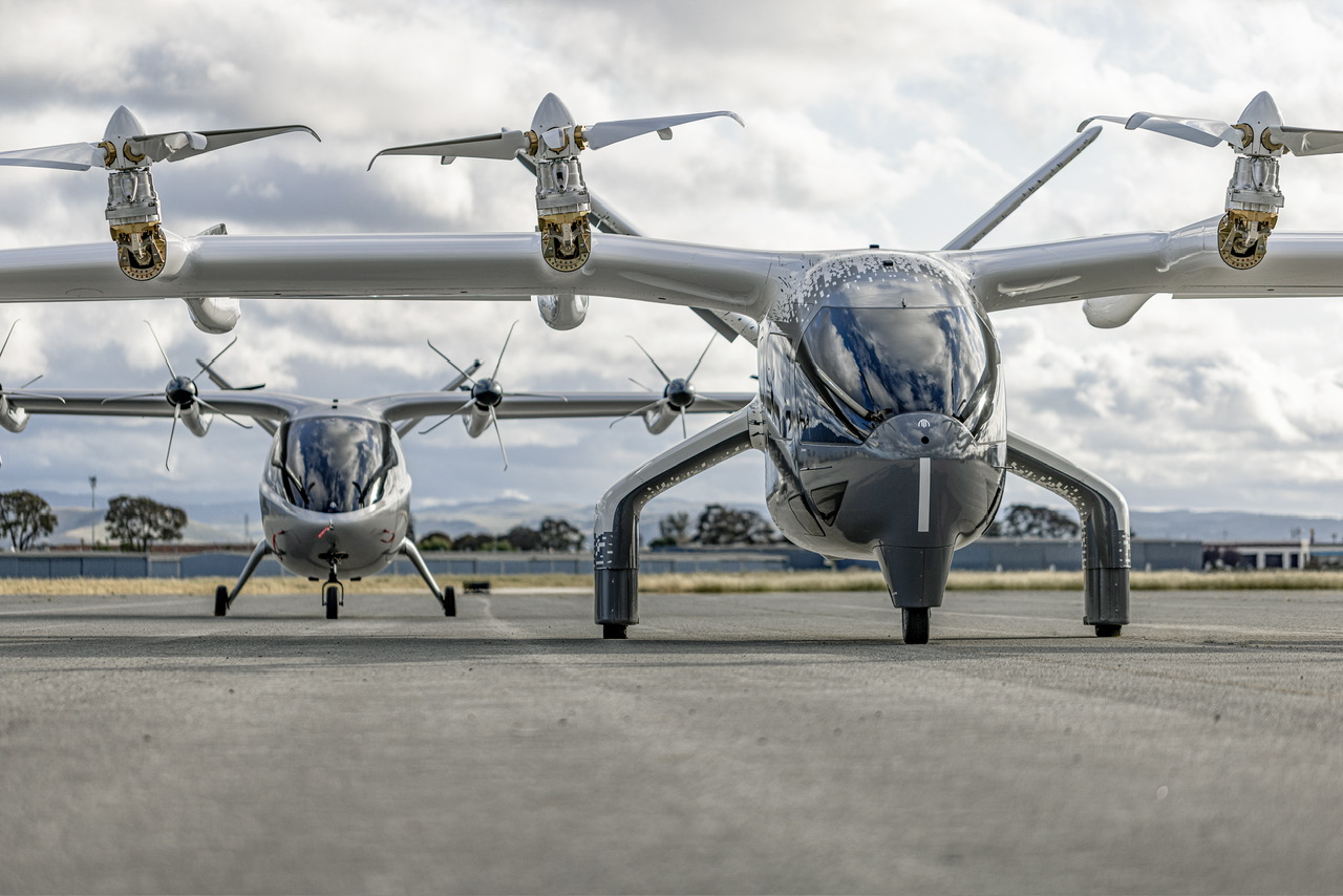 Two black Archer Midnight aircraft sat on a landing strip. A line of tree and a grey sky are visible behind them.
