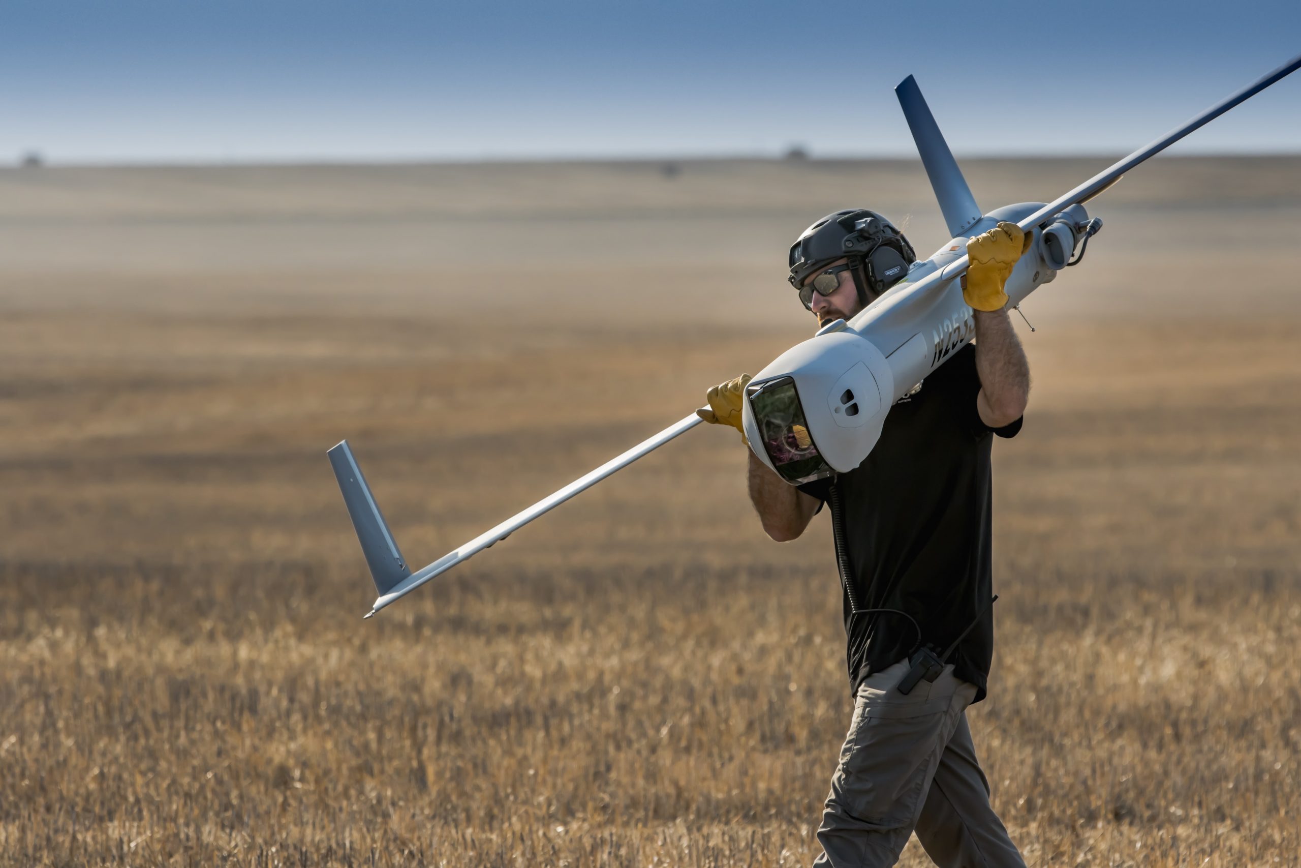 A person dressed in black and wearing safety goggles carries a white insitu drone. The person is walking through a field of wheat in front of a hazy blue sky.