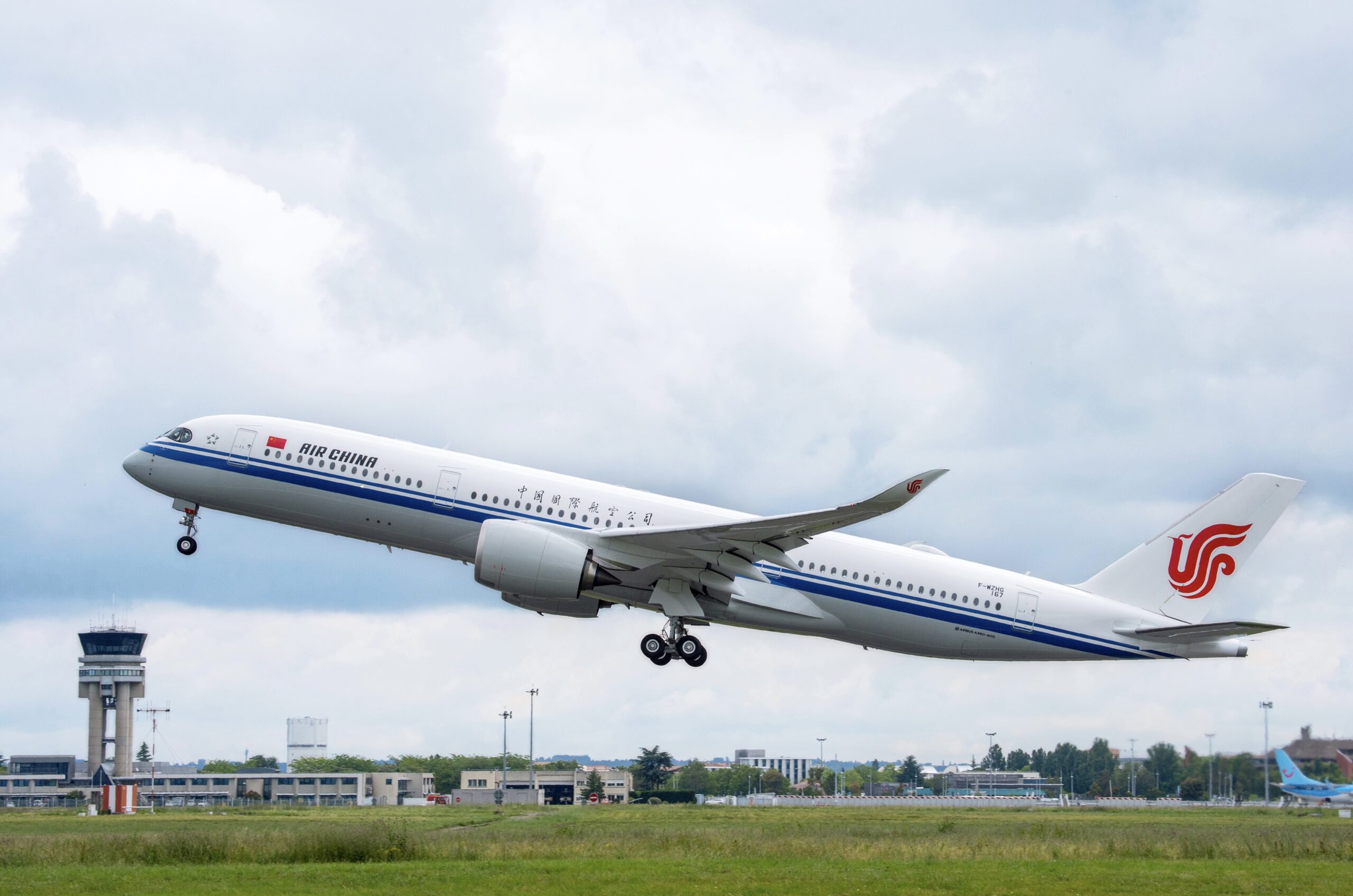 A white, blue, and red Air China jet taking off from an airport. A green field is seen in front of the airport terminal.
