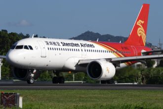 A white, red, and yellow Shenzhen Airlines jet sat on a runway, behind a dark green field.