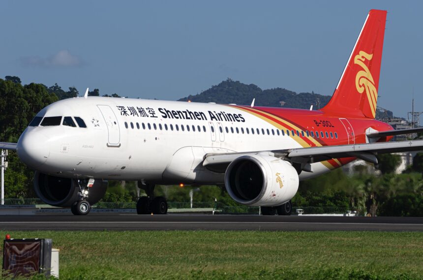 A white, red, and yellow Shenzhen Airlines jet sat on a runway, behind a dark green field.