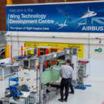 Airbus employees at the Wing Technology Development Centre in Filton, UK. They are seen working on a production line, in front of a blue sign.