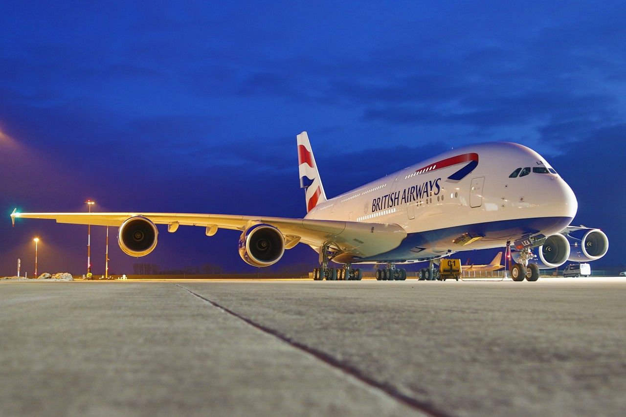 A British Airways Airbus jet sat on a runway at sunset, in front of a dark blue sky.