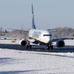 A white, blue, and yellow Ryanair jet on a black runway. The runway is surrounded by snow, and the sky behind is grey.