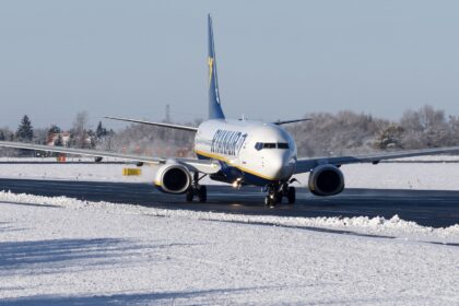 A white, blue, and yellow Ryanair jet on a black runway. The runway is surrounded by snow, and the sky behind is grey.