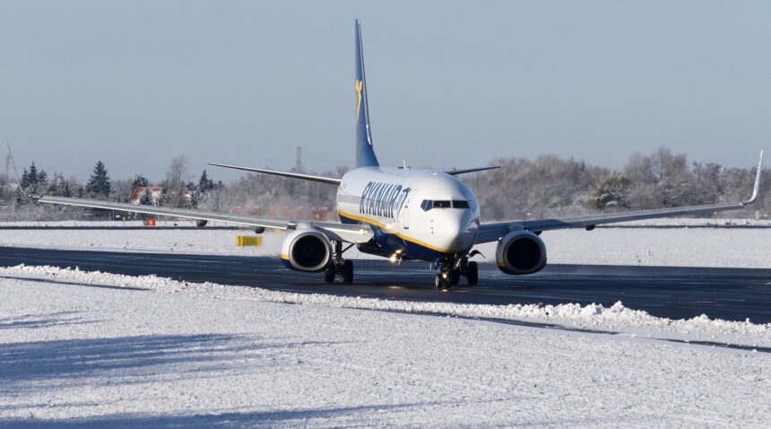 A white, blue, and yellow Ryanair jet on a black runway. The runway is surrounded by snow, and the sky behind is grey.