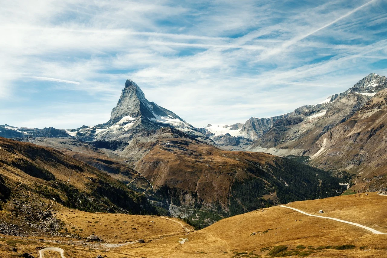 The Matterhorn mountain in the Alps, Switzerland. The surrounding landscape is covered in brown plants, and the sky behind is cloudy.