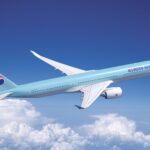 A blue and white Korean Air jet flying upwards in front of a cloudy sky.